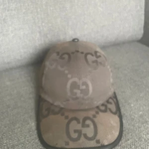 (solbränd) - Säljer en svart keps från Gucci med den klassiska GG-loggan i mörkare toner över hela kepsen. Kepsen har böjd skärm, justerbar rem bak och är tillverkad i bomullsmaterial. Perfekt för dig som vill ha en snygg och lyxig accessoar med streetkänsla. Kepsen är i helhet dunder skick, men väldigt solbränd… Därav det låga priset! (Jumbo heter modellen för de som inte vet)