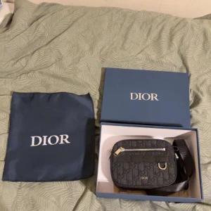 Svart Dior axelväska med monogram - Säljer en svart axelväska från Dior med klassiskt monogrammönster och silverfärgade detaljer. Väskan har en dragkedja framtill, justerbar axelrem och snygga läderdetaljer. Kommer med originalbox och dustbag. Perfekt för dig som vill ha en lyxig och stilren accessoar.