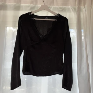 Svart långärmad blus med spets - Snygg svart blus med djup v-ringning och spetsdetaljer längs urringningen. Långärmad och i ett tunt, lätt glansigt material som ger en elegant känsla. 