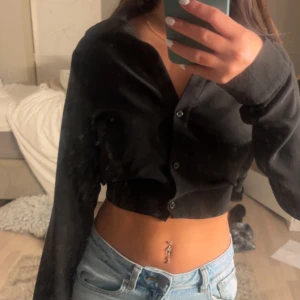 Svart croppad blus med knyt i ryggen - Säljer en svart croppad blus med långa ärmar och knappar framtill. Toppen har snygg knytning i ryggen aldrig använd!
