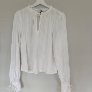 Vit blus med knyt från Divided - Supersöt vit blus från Divided med tunna vertikala ränder i tyget. Blusen har långa ärmar med knyt vid handlederna och en liten volangkant vid halsen. Knytband framtill vid halsringningen ger en extra detalj. Perfekt för en chill och stilren look. Endast använd 1 gång, då jag behövde något akut att jobba i!