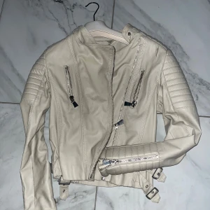 Beige skinnjacka med dragkedjor - Snygg beige skinnjacka med coola detaljer som silvriga dragkedjor, quiltade partier på axlar och ärmar samt bälten vid midjan. Jackan har en asymmetrisk dragkedja framtill och flera fickor med dragkedja. Perfekt för dig som vill ha en edgy look.
