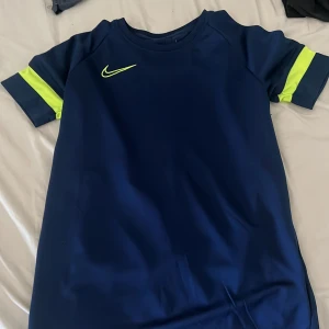 Marinblå Nike fotbollströja - Snygg marinblå fotbollströja från Nike med korta ärmar och neongula detaljer på ärmarna samt Nike-logga på bröstet. Tillverkad i ett lätt och funktionellt material som passar perfekt för träning och match.