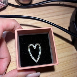 Silverring med glittrigt hjärta - Säljer en snygg silverfärgad ring med ett öppet hjärta som är täckt av glittrande stenar. Ringen har en modern och unik design där hjärtat är lite utdraget och ger en cool vibe. Perfekt för dig som vill sticka ut med dina accessoarer. Justerbar tror den är köpt på glitter eller guldfynd 
