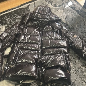 Svart glansig dunjacka från Moncler Maya - Säljer en svart, glansig dunjacka från Moncler med klassisk pufferstil. Jackan har huva, två stora fickor med dragkedja och Moncler-logga på ärmen. Perfekt för kalla dagar och riktigt snygg till streetwear-looken.