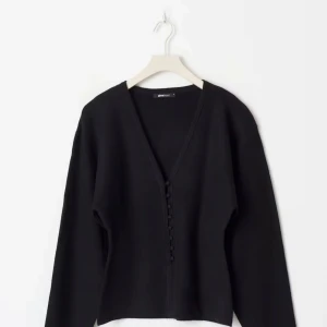 Svart kofta med knappar från Gina Tricot - Svart kofta med djup v-ringning och snygga klädda knappar framtill. Långärmad och i mjukt stickat material som känns skönt mot huden. Perfekt till både jeans och kostymbyxor för en clean look. Aldrig använd 