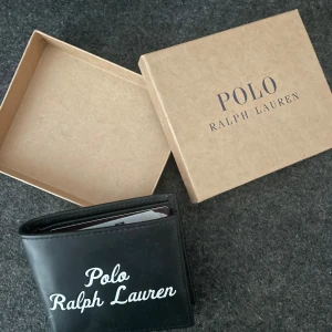 Plånbok från Ralph Lauren - Snygg svart plånbok från Polo Ralph Lauren i stilren design. Tillverkad i skinn med vit logotyp på framsidan. Perfekt storlek för kort och sedlar, levereras med original ask. Köptes i Juli i Paris, Galeries LaFayette för 120 Euro. Säljer den nu för 480kr. Pris går att diskuteras.