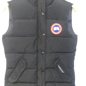 Svart dunväst från Canada Goose XS - Svart dunväst från Canada Goose med hög krage och klassisk logga på bröstet. Västen har två snedställda fickor med knappar och stängs med både dragkedja och tryckknappar. Perfekt för lager-på-lager under kyliga dagar. Obs den är  ny! 