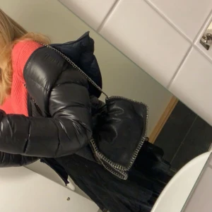 Svart axelväska med kedjedetaljer - Säljer en svart axelväska från liknande Stella McCartney med snygga silverfärgade kedjor längs kanterna och som axelrem. Väskan har en mjuk form och är gjord i syntetmaterial som liknar mocka. Perfekt för dig som vill ha en cool och trendig väska med statement-detaljer.