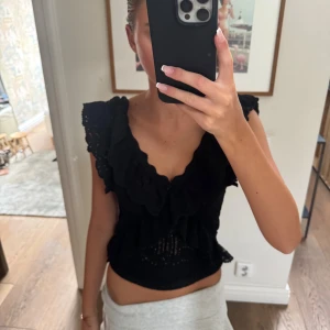 Svart volangblus med öppen rygg - Supersöt svart blus med volangdetaljer och broderade kanter. Djup v-ringning fram och öppen rygg ger en unik look. Ärmlös modell med croppad passform, perfekt till höga jeans eller kjol. Luftigt material som känns lätt på kroppen.