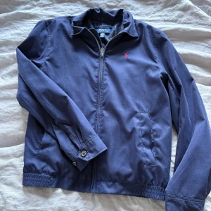 Polo Ralph Lauren jacka  - Snygg marinblå jacka från Polo Ralph Lauren med dragkedja framtill och rutigt innerfoder. Perfekt för dig som gillar klassisk och clean stil. Storlek 14-16 år L juniorstorlek 