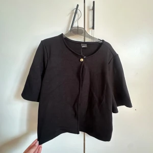 Svart bolerotopp från Gina Tricot - Svart bolerotopp från Gina Tricot med rund halsringning och en dekorativ guldig knapp framtill. Perfekt att slänga över en klänning eller topp för en snygg layering-effekt.