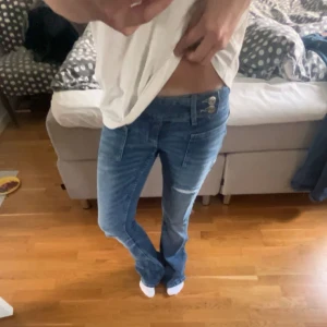 Blå bootcut jeans med slitningar - Snygga blå bootcut jeans med slitningar på benen och framsydda fickor. Jeansen har låg midja och klassisk knappstängning med två synliga knappar. Perfekta för en avslappnad och trendig look.