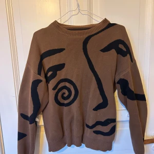 Brun stickad tröja med ansiktsmönster - Cool brun stickad tröja från Sweet SKTBS med svart abstrakt ansiktsmönster över hela framsidan och ärmarna. Tröjan har rund halsringning och ribbade muddar. Perfekt för dig som gillar unika statement-plagg.