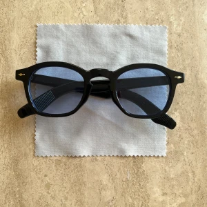 Svarta glasögon med blåtonade linser - Säljer ett par svarta glasögon med tjock, rundad båge i acetat och blåtonade linser. Klassisk retrostil med små detaljer på skalmarna. Perfekt för dig som vill ha en cool och tidlös look.