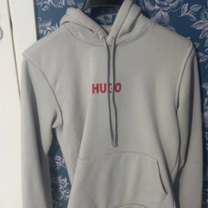 Ljusgrå hoodie från HUGO med röd logga - Snygg ljusgrå hoodie från HUGO med stor röd logga både fram och bak. Klassisk modell med huva och dragsnören samt en stor magficka. Perfekt för dig som gillar stilrena plagg med tydlig branding. Mjuk och skön bomullsblandning.