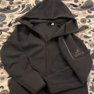 Svart hoodie från Adidas med dragkedja - Svart hoodie från Adidas med hel dragkedja och huva. Tröjan har Adidas-logga på ärmen och en diskret grå detalj längs axeln. Skön passform och perfekt för chill eller träning. Materialet är mjukt och känns som polyester eller fleece.