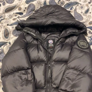Svart pufferjacka från Canada Goose - Säljer en svart pufferjacka från Canada Goose i storlek M. Jackan har en stor huva, tydlig logga på ärmen och dragkedja framtill. Den är riktigt fluffig och perfekt för kalla dagar. Klassisk design med diskreta detaljer och snygg passform. Jag köpte den begagnad för 7000 med prislapp. Säljer den nu för 4000