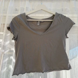 Grå croppad topp från H&M Divided - Enkel och snygg grå croppad topp från H&M Divided med v-ringning och korta ärmar. Toppen har vågig kant nertill och är tillverkad i mjuk bomull, perfekt för en chill och stilren look.