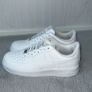 Nike Air Force 1 helvita sneakers - Klassiska Nike Air Force 1 sneakers i helvitt skinn med perforerad tå, vit sula och snörning. Snygg och clean siluett med rund tå och diskret Nike-logga på sidan och hälen. Perfekta för dig som gillar en stilren look.