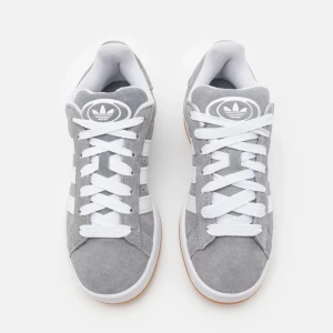 Grå adidas Gazelle sneakers mocka - Snygga adidas Gazelle sneakers i grå mocka med vita detaljer och klassiska tre ränder på sidorna. Skorna har vit sula, rund tå och snörning. Perfekta för dig som gillar en clean och sportig stil.