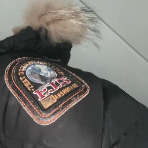 Svart Parajumpers dunjacka med päls - Säljer en svart Parajumpers dunjacka med ikonisk gul rem och stor pälsluva. Jackan har patch på ärmen, dragkedja och flera fickor. Slim fit-modell som håller dig varm och snygg hela vintern. Perfekt för dig som vill ha en klassisk och trendig vinterjacka.Äkta