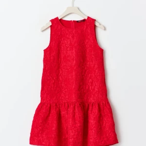 Röd ärmlös kortklänning med struktur - Supersnygg röd kortklänning med strukturerat tyg och volang nedtill. Klänningen är ärmlös och har en rund halsringning. Perfekt för dig som vill sticka ut med en färgstark look. Lätt att styla med både sneakers och klackar.