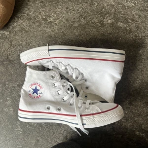 Vita Converse All Star high tops - Klassiska vita Converse All Star high tops med röd och blå rand på sulan. Skorna har snörning, rund tå och är tillverkade i canvas. Perfekta för en avslappnad och cool stil. Converse-logga på sidan och bak på hälen.