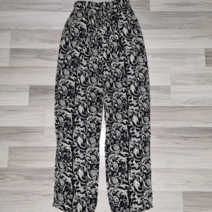 Svarta byxor med blommigt mönster - Snygga svarta byxor med vitt blommigt mönster och elastisk midja. Lätta och luftiga, perfekta för dig som gillar en avslappnad stil. Mjukt material som känns skönt mot huden och ger en cool vibe till outfiten. Sömmen har gått upp i vänster sida av midjan,  lätt att fixa.