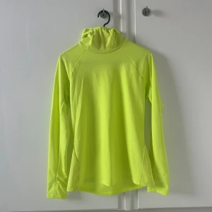 Neongul långärmad träningströja - Neongul långärmad träningströja med hög krage och tumhål vid ärmsluten. Tillverkad i ett lätt och ventilerande syntetmaterial som passar perfekt för löpning eller annan träning. Enkel och sportig design som verkligen syns. Storlek S. Aldrig använd.
