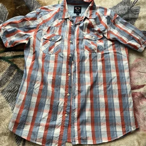 Rutig kortärmad skjorta från J.F.K - Kortärmad skjorta från J.F.K i storlek XL med rött, blått och vitt rutmönster. Skjortan har två bröstfickor med lock och knappar, klassisk krage och knäppning framtill. Perfekt för en avslappnad och cool stil. Vid frågor hör av dig!