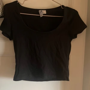 Svart croppad topp från H&M Divided - En svart croppad topp från H&M Divided med rundad, djup urringning och korta ärmar. Enkel och stilren design i mjuk bomull, perfekt att matcha med jeans eller kjol för en clean look.