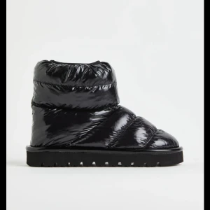 Svarta uggs i glansigt material - Säljer ett par svarta moon boots med glansig yta och vadderad design. Skorna har en chunky sula med grovt mönster och är perfekta för kalla dagar. De är tillverkade i syntetmaterial och har en modern, puffig look som sticker ut.