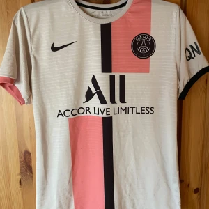 PSG Messi 30 bortatröja Nike S - Paris Saint-Germain bortatröja med Messi 30-tryck. Tröjan är vit med svarta och rosa vertikala ränder, PSG-märke på bröstet och Nike-logga. Tillverkad i lätt och ventilerande Dri-FIT-material, kortärmad och med rund hals. Står L på tröjan men sitter som en s