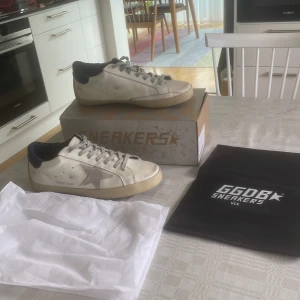 Golden Goose Superstar sneakers vita - Golden Goose Superstar sneakers i storlek 45 funkar för 44, vitt skinn med grå stjärndetalj på sidan och mörkblå hälkappa. Klassisk låg modell med snörning och beige sula. Snygg och avslappnad look, perfekt till jeans eller shorts. Kommer med originalkartong och dustbag. 
