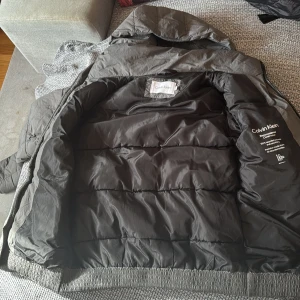 Svart pufferjacka från Calvin Klein XS - Säljer en svart pufferjacka från Calvin Klein i storlek XS. Jackan har huva, dragkedja framtill och är tillverkad i 100% återvunnen polyester. Snygg och enkel design med diskret logga på ärmen och två fickor framtill. Original pris 3,500kr