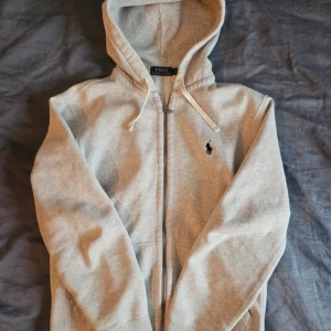 Ralph lauren zip hoodie - Snygg ljusgrå hoodie från Polo Ralph Lauren med dragkedja och huva. Tröjan har snörning i huvan, klassisk logga broderad på bröstet och är tillverkad i mjuk bomull. Perfekt för chill dagar eller när du vill ha en clean och enkel look.