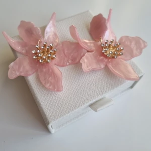 Stora rosa blomörhängen med strass - Superfina statement-örhängen i form av stora blommor i ljusrosa plast med glittrande strassdetaljer i mitten och guldfärgade inslag. Perfekta för dig som vill sticka ut och addera en söt touch till din look. Lådan ingår ej.