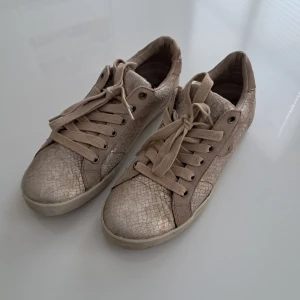 Beige sneakers från Tamaris, stl 40 - Snygga beige sneakers från Tamaris med ormskinnsmönstrad yta och klassisk snörning. Skorna har rund tå och platt sula, vilket gör dem riktigt bekväma. Materialet är läder med detaljer i mockaimitation. Perfekta för dig som gillar en stilren men ändå unik look. Tvättade! 