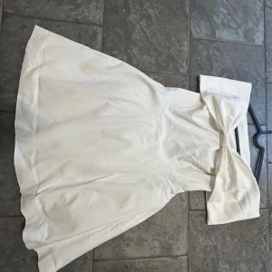 Vit offshoulder klänning från NA-KD - Superfin vit kortklänning från NA-KD med offshoulder-modell och stor rosettdetalj framtill. Klänningen har en figurnära passform upptill och utställd kjol. Perfekt till studenten! Aldrig använd! Nypris 800kr.