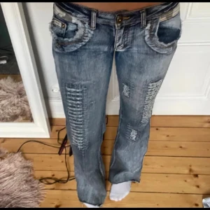Blå slitna bootcut jeans - Snygga blå jeans med slitningar och råa detaljer på fickor och ben. Modellen har bootcut och låg midja, med klassisk knappstängning och coola sömmar. Perfekta för en avslappnad streetstil.                                        Midjemått: 38 cm rakt över                                                 Innerbenslängden: 80 cm 