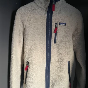 Beige fleecejacka från Patagonia - Säljer en mysig beige fleecejacka från Patagonia med dragkedja i kontrasterande mörkblå färg. Jackan har tre fickor med dragkedjor och röda detaljer. Perfekt för kyliga dagar! Den kommer att sitta perfekt för dig som är runt 170-175. 