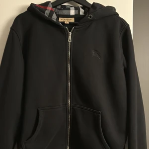 Svart hoodie från Burberry med dragkedja - Svart hoodie från Burberry med klassiskt rutigt foder i huvan och broderad logga på bröstet. Tröjan har dragkedja framtill, två fickor och en skön huva. Perfekt för dig som vill ha en stilren och lyxig look. Dock har jag inga snören till