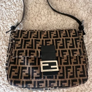 Fendi zucca canvas baguette 1990s/2000s - Säljer en ikonisk Fendi axelväska i brunt och svart med klassiskt FF-logomönster över hela väskan. Väskan har justerbar axelrem i svart skinn och silverfärgat spänne framtill med Fendi-gravyr. Materialet är canvas med detaljer i skinn.