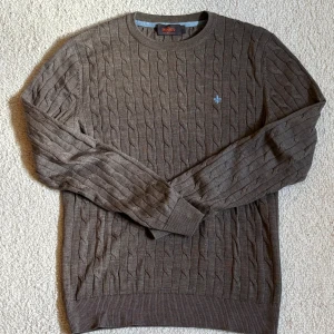 Brun kabelstickad tröja från Morris - Kabelstickad brun tröja från Morris i 100% merinoull. Rund halsringning, ribbade muddar och diskret ljusblå broderad logga på bröstet. Perfekt för lager-på-lager och snygg till jeans eller chinos.