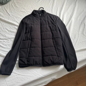 Svart pufferjacka med dragkedja - Stilren svart pufferjacka med quiltade paneler framtill och släta ärmar. Jackan har hög krage, hel dragkedja och två sidofickor. Perfekt för kyligare dagar och enkel att matcha med olika outfits.