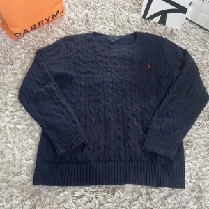 Mörkblå kabelstickad tröja Polo Ralph Lauren - Kabelstickad mörkblå tröja från Polo Ralph Lauren med klassisk rund halsringning och röd broderad logga på bröstet. Tröjan har långa ärmar och breda ribbade muddar vid ärmslut och nederkant. Perfekt för en stilren och chill look.