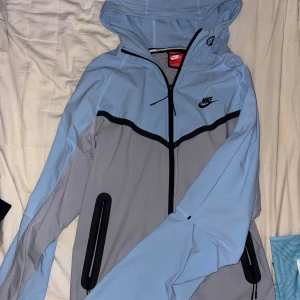 Grå tracksuit från Nike med dragkedjor - Snygga grå tracksuit från Nike med svarta dragkedjor på fickorna och snörning i midjan. Byxorna har en sportig look och är tillverkade i ett lätt syntetmaterial som passar perfekt för träning. Nike-logga på låret och detaljer vid benslut. Storleken är S i tröjan och M i byxorna men skulle säga det passar M-L då det är en pösigare variant av tracksuit
