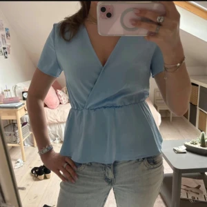 Ljusblå omlottopp från Vila - Supersöt ljusblå omlottopp från Vila med korta ärmar och v-ringning. Toppen har resår i midjan som ger en snygg peplum-effekt och är tillverkad i ett mjukt, lätt material. Perfekt för dig som gillar en feminin och fräsch stil.