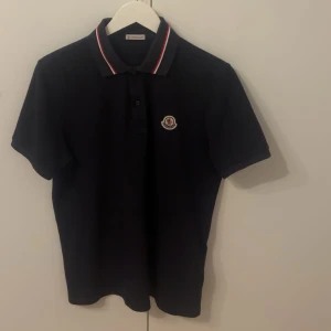 Mörkblå Moncler pikétröja med logga - Svart pikétröja från Moncler med klassisk krage och rödvit rand längs kragen. Broderad Moncler-logga på bröstet och diskret Moncler-tryck i nacken. Tillverkad i bomull för en skön och stilren känsla.